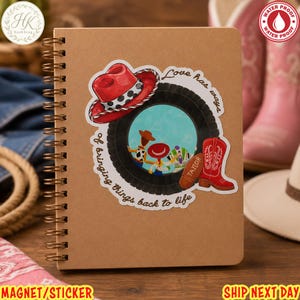 May include: A spiral-bound notebook with a tan cover featuring a sticker of a black tire framing a cartoon scene with a red cowboy hat, a red boot labeled Taylor, and the text Love has ways of bringing things back to life in black script.