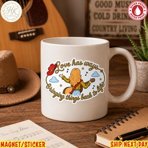 May include: A white ceramic mug featuring a graphic illustration of a person with long blonde hair playing an acoustic guitar. The design includes a red cowboy hat, blue clouds, musical notes, and the text Love has ways, Bringing things back to life.