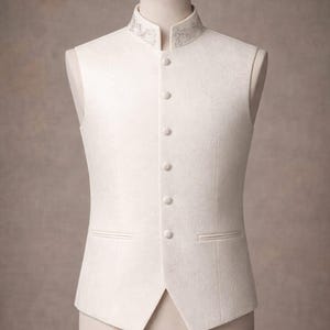 May include: Ivory-colored formal vest with a mandarin collar and decorative embroidery. The vest features a six-button closure, two front pockets, and a tailored fit. Displayed on a mannequin, the vest is suitable for weddings or formal events.