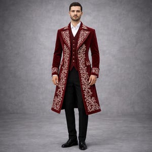 Puede incluir: Un abrigo de terciopelo burdeos con bordados dorados. El abrigo presenta un diseño largo y entallado con un chaleco a juego, combinado con pantalones negros y zapatos de vestir. El conjunto es formal y elegante.