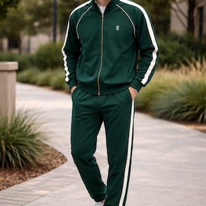 Puede incluir: Un chándal verde oscuro con rayas y detalles en blanco. La chaqueta tiene cierre de cremallera y un pequeño logotipo en el pecho. Los pantalones a juego tienen una raya blanca en el lateral. La persona lleva zapatillas blancas.
