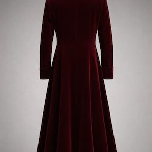 May include: A long, burgundy velvet coat with a high collar and long sleeves. The coat has a flared skirt and is displayed against a neutral background. The fabric appears soft and luxurious.