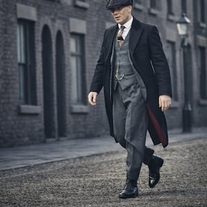 Puede incluir: Un hombre con vestimenta formal, que incluye un abrigo largo negro con forro rojo, un chaleco gris y pantalones a juego. Lleva una gorra plana y botas negras. La escena tiene lugar en una calle adoquinada.