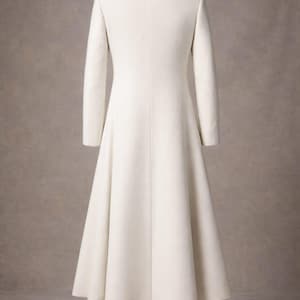 May include: A long, ivory-colored coat with a high collar and long sleeves. The coat has a flared skirt and is displayed on a mannequin. The collar has a decorative design. The mannequin is wearing silver boots.