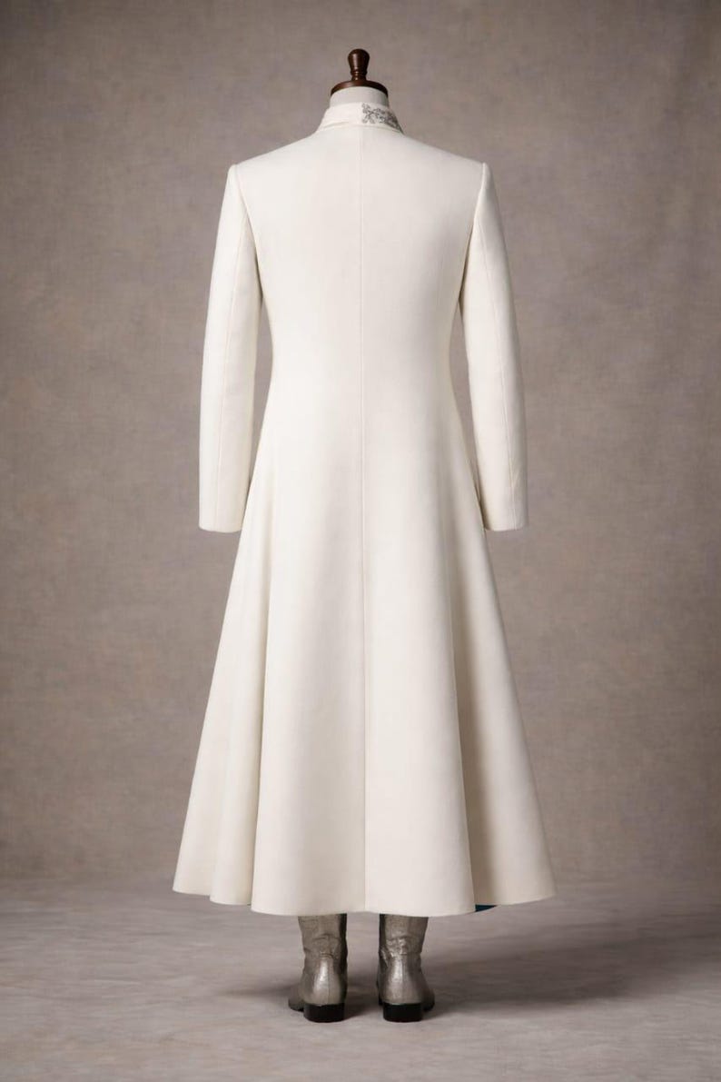 May include: A long, ivory-colored coat with a high collar and long sleeves. The coat has a flared skirt and is displayed on a mannequin. The collar has a decorative design. The mannequin is wearing silver boots.