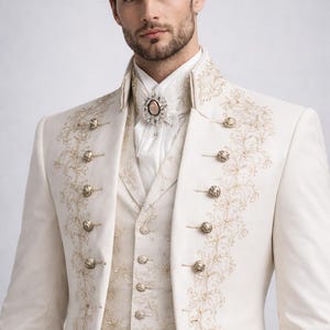 May include: Ivory-colored formal suit with gold embroidery and ornate buttons. The suit includes a double-breasted jacket, vest, and high-collared shirt. A decorative brooch adorns the shirt. The suit is suitable for weddings or formal events.