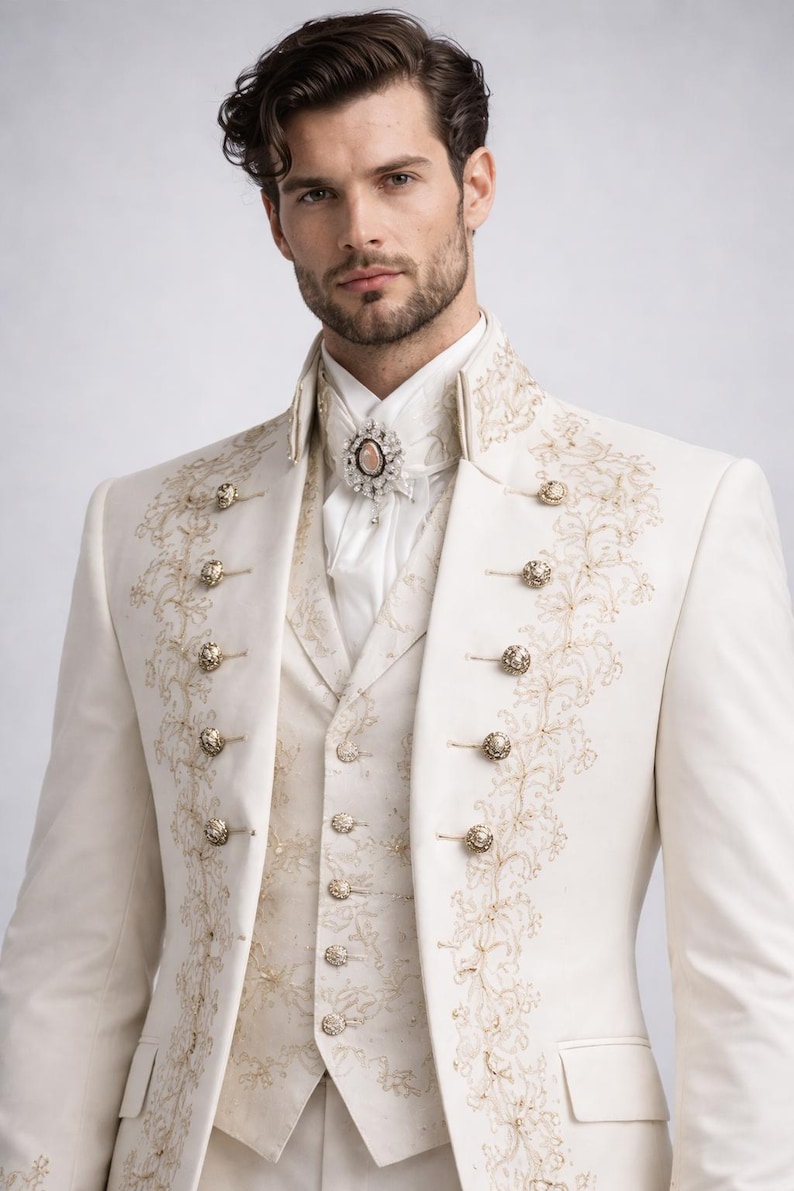 May include: Ivory-colored formal suit with gold embroidery and ornate buttons. The suit includes a double-breasted jacket, vest, and high-collared shirt. A decorative brooch adorns the shirt. The suit is suitable for weddings or formal events.