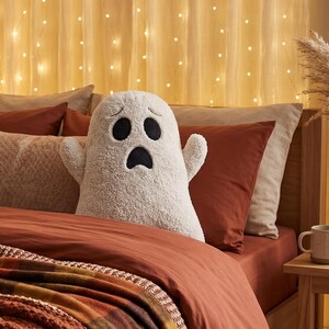 Pode incluir: Uma almofada de pel&uacute;cia em forma de fantasma, na cor branco-sujo, com uma superf&iacute;cie texturizada semelhante a l&atilde; de carneiro. O fantasma apresenta olhos ovais pretos bordados e uma boca preta com express&atilde;o de surpresa, com dois bra&ccedil;os.