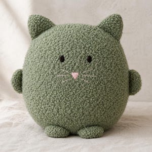 May include: A round, olive green plush cat toy with a textured, nubby fabric surface. It features two pointed ears, small black embroidered eyes, a light pink triangular nose, and thin white embroidered whiskers on its circular body.
