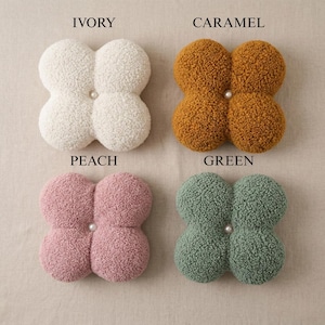 May include: Four plush, clover-shaped decorative cushions in ivory, caramel, peach, and green. Each cushion features a central pearl-like button. The cushions are made of a soft, textured material.