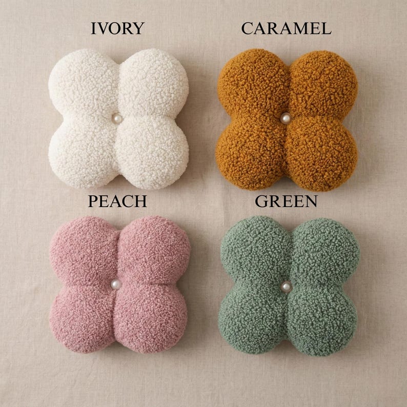 May include: Four plush, clover-shaped decorative cushions in ivory, caramel, peach, and green. Each cushion features a central pearl-like button. The cushions are made of a soft, textured material.