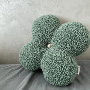 May include: A sage green, four-lobed decorative cushion with a textured, plush surface. A pearl-like button secures the lobes at the centre. The cushion rests on a grey surface, with a textured grey wall in the background.
