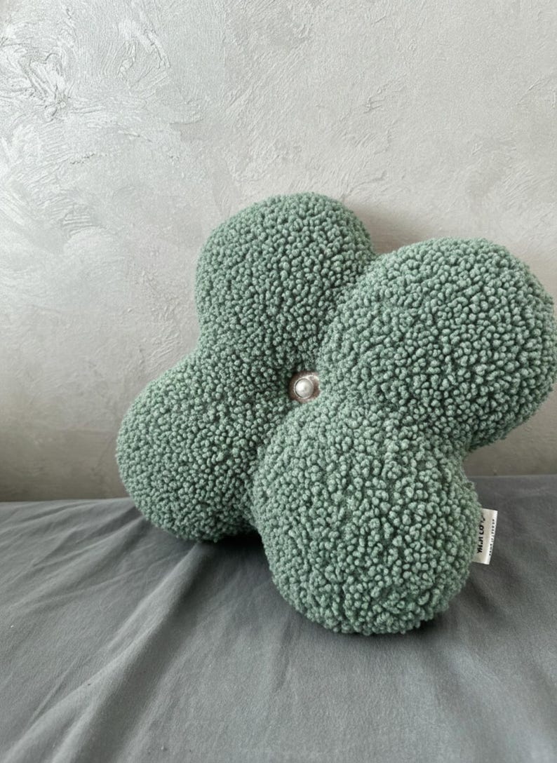 May include: A sage green, four-lobed decorative cushion with a textured, plush surface. A pearl-like button secures the lobes at the centre. The cushion rests on a grey surface, with a textured grey wall in the background.