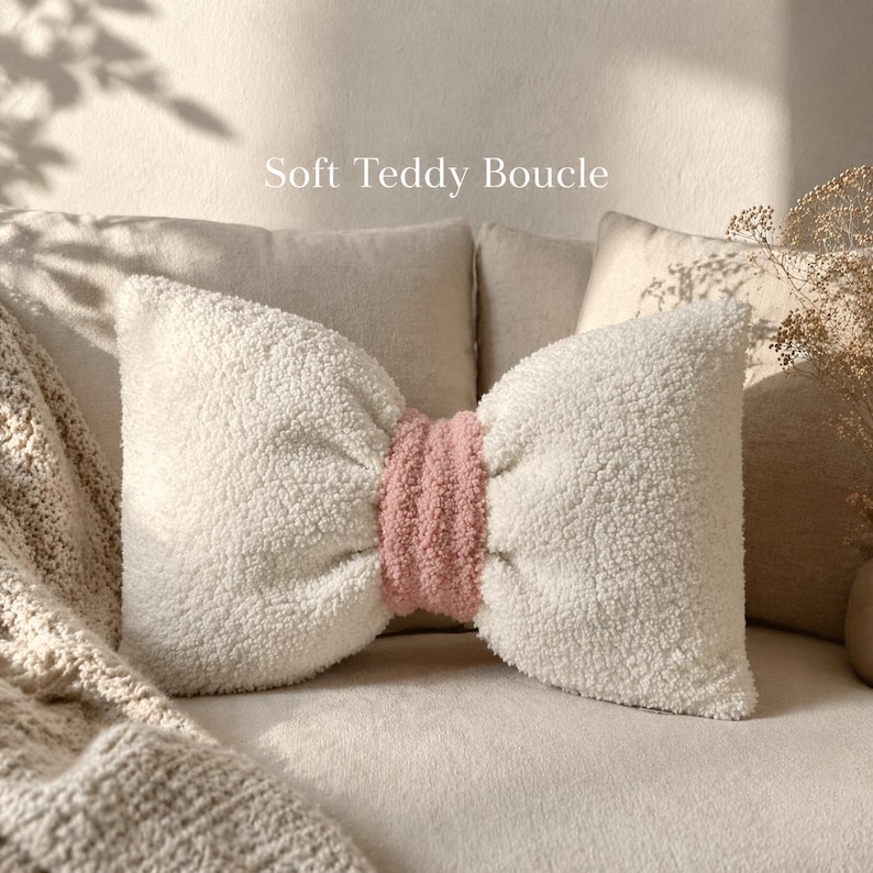 May include: A soft, bow-shaped pillow with a white teddy boucle texture and a pink center. The pillow is placed on a beige couch with a matching blanket. The text "Soft Teddy Boucle" is displayed on the image.