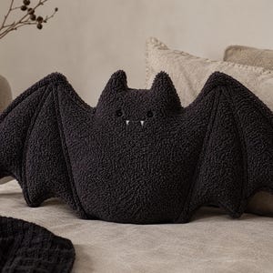 May include: A dark charcoal grey bat-shaped throw cushion made of textured, curly fabric. The cushion features two pointed ears, wide outstretched wings, and a small face with two embroidered eyes and two tiny white fangs visible at the mouth.