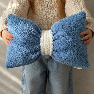 May include: A large, bow-shaped pillow in shades of blue and white. The pillow has a textured, plush appearance, with a light blue bow and a white center. The pillow is held up by a person.