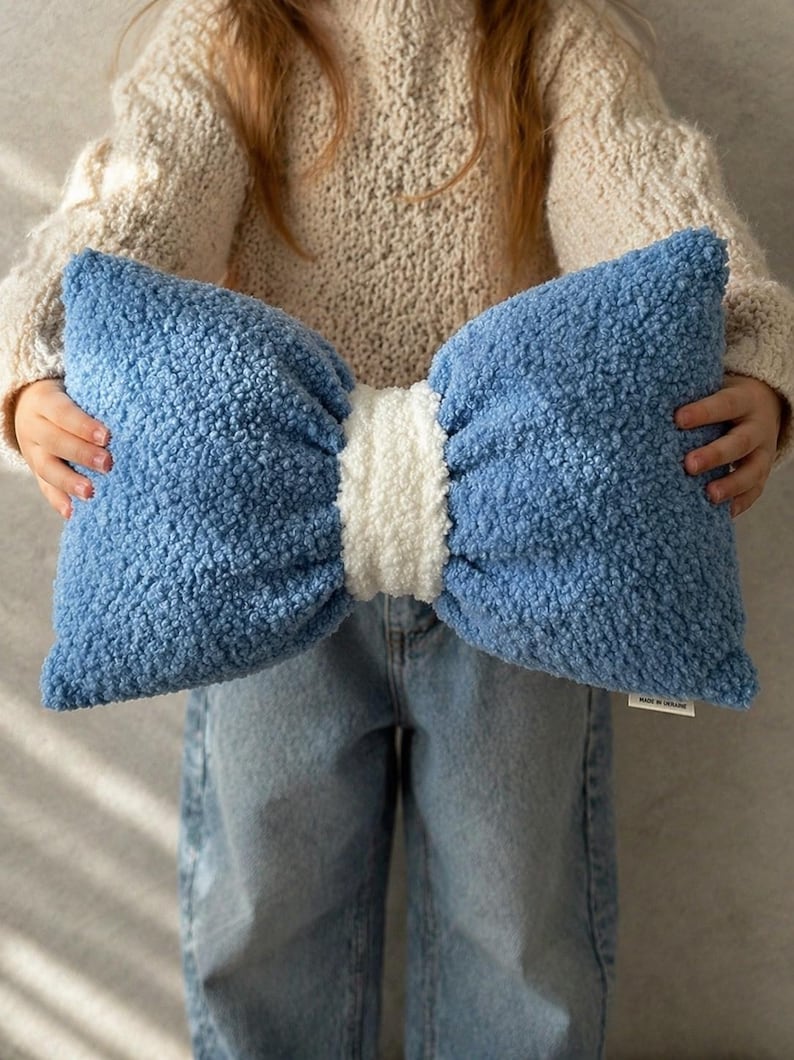 May include: A large, bow-shaped pillow in shades of blue and white. The pillow has a textured, plush appearance, with a light blue bow and a white center. The pillow is held up by a person.