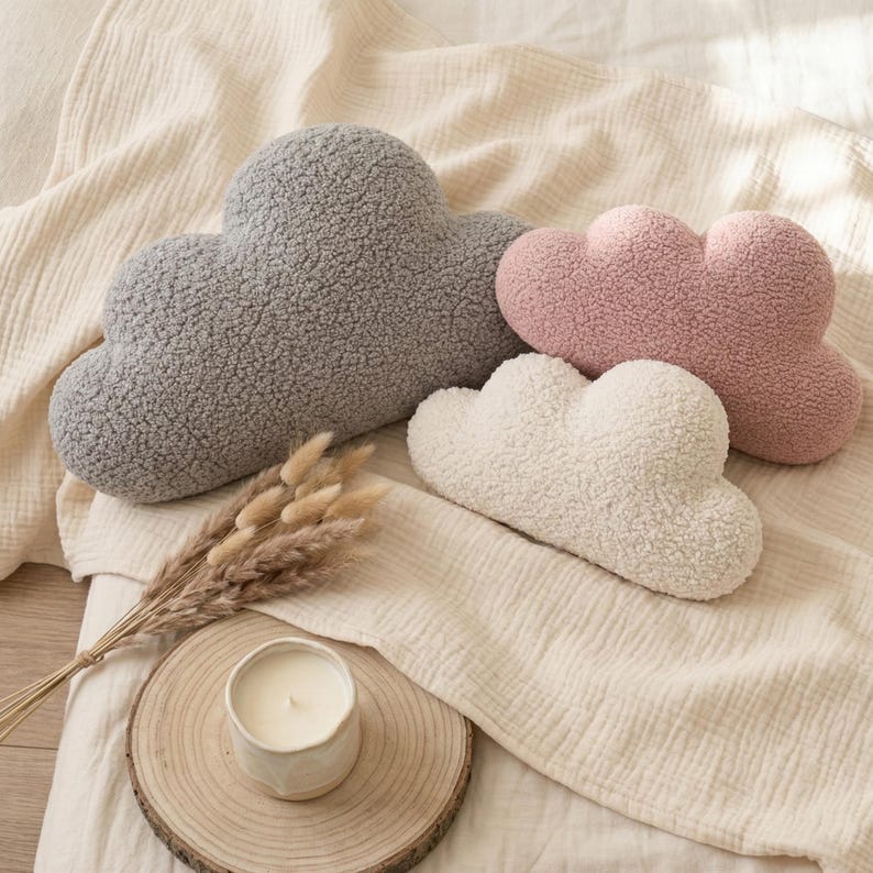 May include: Four cloud-shaped decorative pillows in gray, pink, and white, with a textured, fluffy appearance. The pillows are arranged on a cream-colored fabric. A candle in a white ceramic holder sits on a wooden slice.