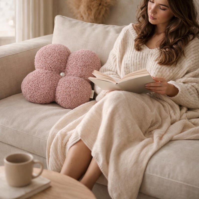 May include: A flower-shaped, dusky pink pillow with a pearl-like centre rests on a beige sofa. The pillow has a textured, plush appearance. A person is reading a book while wrapped in a cream-coloured blanket and sweater.