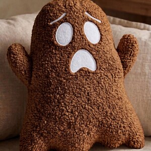 Pode incluir: Uma almofada em forma de fantasma castanha feita de tecido polar texturizado e encaracolado. A almofada apresenta dois olhos ovais brancos e uma express&atilde;o de boca aberta branca cosidos na frente, com dois pequenos bra&ccedil;os nas laterais.