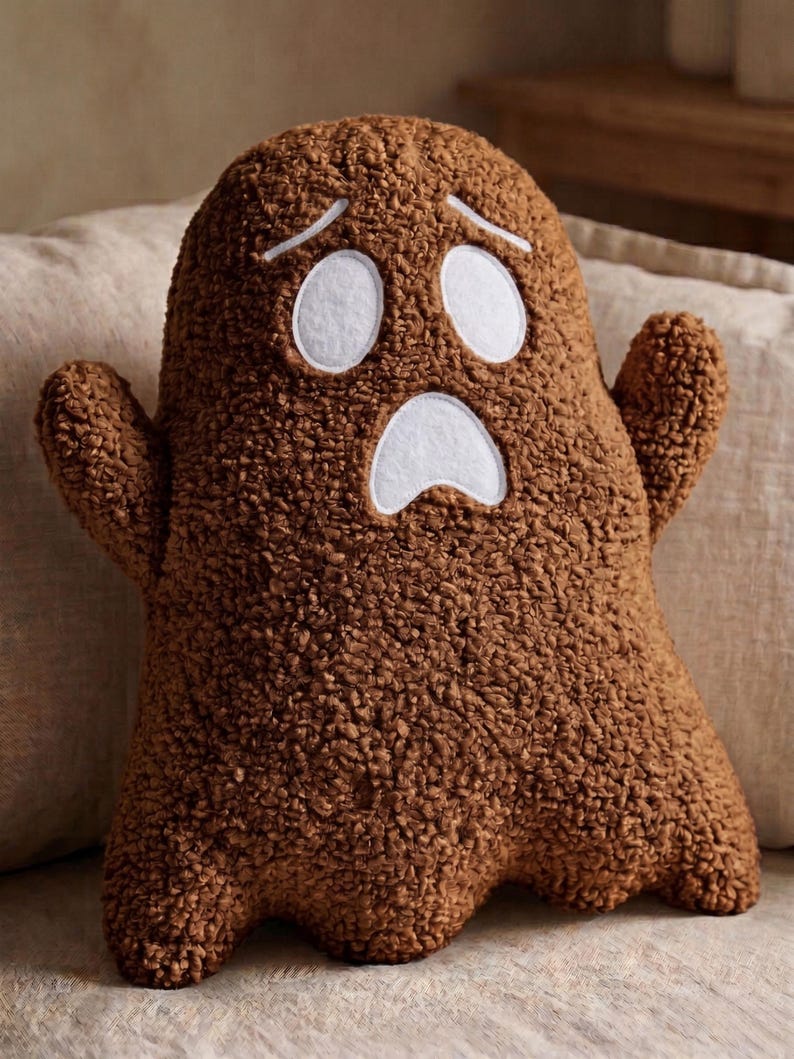 Pode incluir: Uma almofada em forma de fantasma castanha feita de tecido polar texturizado e encaracolado. A almofada apresenta dois olhos ovais brancos e uma express&atilde;o de boca aberta branca cosidos na frente, com dois pequenos bra&ccedil;os nas laterais.