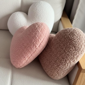 Peut inclure: Trois coussins en forme de cœur, dans des tons de rose et de blanc. Les coussins ont un aspect doux, texturé et moelleux. Ils sont disposés sur une surface claire et texturée, suggérant un thème de décoration intérieure cosy.