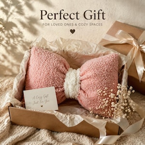 May include: A pink bow-shaped pillow with a white center, presented in a cardboard box with a gift card that reads "A Cozy Gift Just For You." The image includes the text "Perfect Gift" and "FOR LOVED ONES & COZY SPACES."