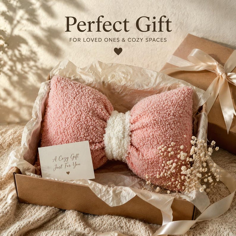 May include: A pink bow-shaped pillow with a white center, presented in a cardboard box with a gift card that reads "A Cozy Gift Just For You." The image includes the text "Perfect Gift" and "FOR LOVED ONES & COZY SPACES."