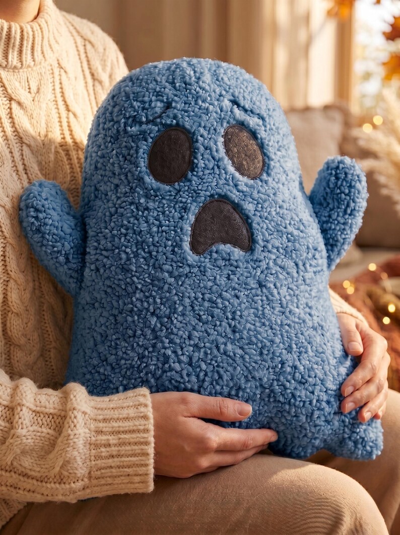 Pode incluir: Uma almofada de pel&uacute;cia azul em forma de fantasma feita de tecido sherpa texturizado. A almofada apresenta dois olhos ovais cinza-escuros e uma boca triangular cinza-escura, dando-lhe uma express&atilde;o de surpresa. Tem dois pequenos bra&ccedil;os laterais.