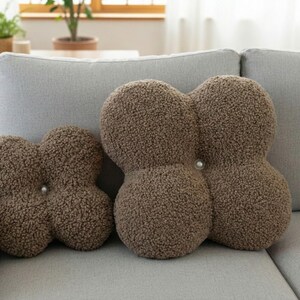 May include: Three brown, textured decorative cushions in a cloverleaf shape sit on a grey sofa. The cushions have a soft, plush appearance and are accented with a single button in the centre of each cushion.