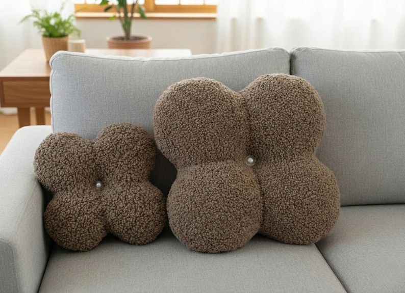 May include: Three brown, textured decorative cushions in a cloverleaf shape sit on a grey sofa. The cushions have a soft, plush appearance and are accented with a single button in the centre of each cushion.