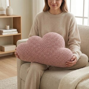May include: A cloud-shaped decorative pillow in a soft, textured, dusty rose color. The pillow is held by a person sitting on a beige sofa. The pillow's fluffy texture is visible, suggesting a cozy and comfortable design. The overall aesthetic is soft and inviting.