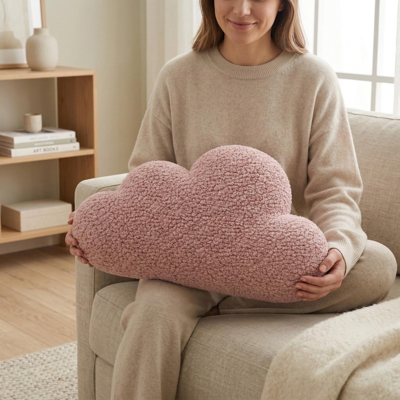 May include: A cloud-shaped decorative pillow in a soft, textured, dusty rose color. The pillow is held by a person sitting on a beige sofa. The pillow's fluffy texture is visible, suggesting a cozy and comfortable design. The overall aesthetic is soft and inviting.