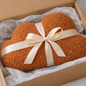 May include: A cloud-shaped, textured, orange pillow tied with a cream-colored ribbon bow, presented in a brown cardboard box with white tissue paper. The pillow is a warm, inviting color.