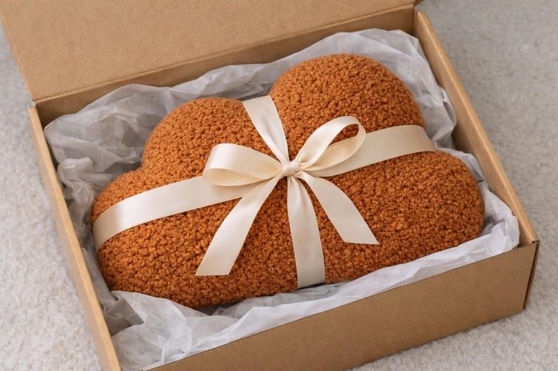 May include: A cloud-shaped, textured, orange pillow tied with a cream-colored ribbon bow, presented in a brown cardboard box with white tissue paper. The pillow is a warm, inviting color.