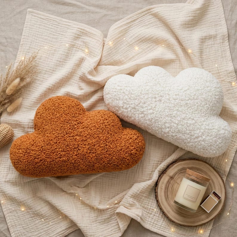May include: Two cloud-shaped decorative pillows, one in a warm orange-brown and the other in white, rest on a beige fabric. The pillows have a textured, fluffy appearance. A small candle in a glass jar sits on a wooden coaster.