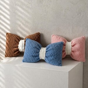 May include: Three bow-shaped decorative pillows in brown, blue, and pink. Each pillow has a white center and a textured, plush appearance. The pillows are arranged on a white surface against a neutral background.
