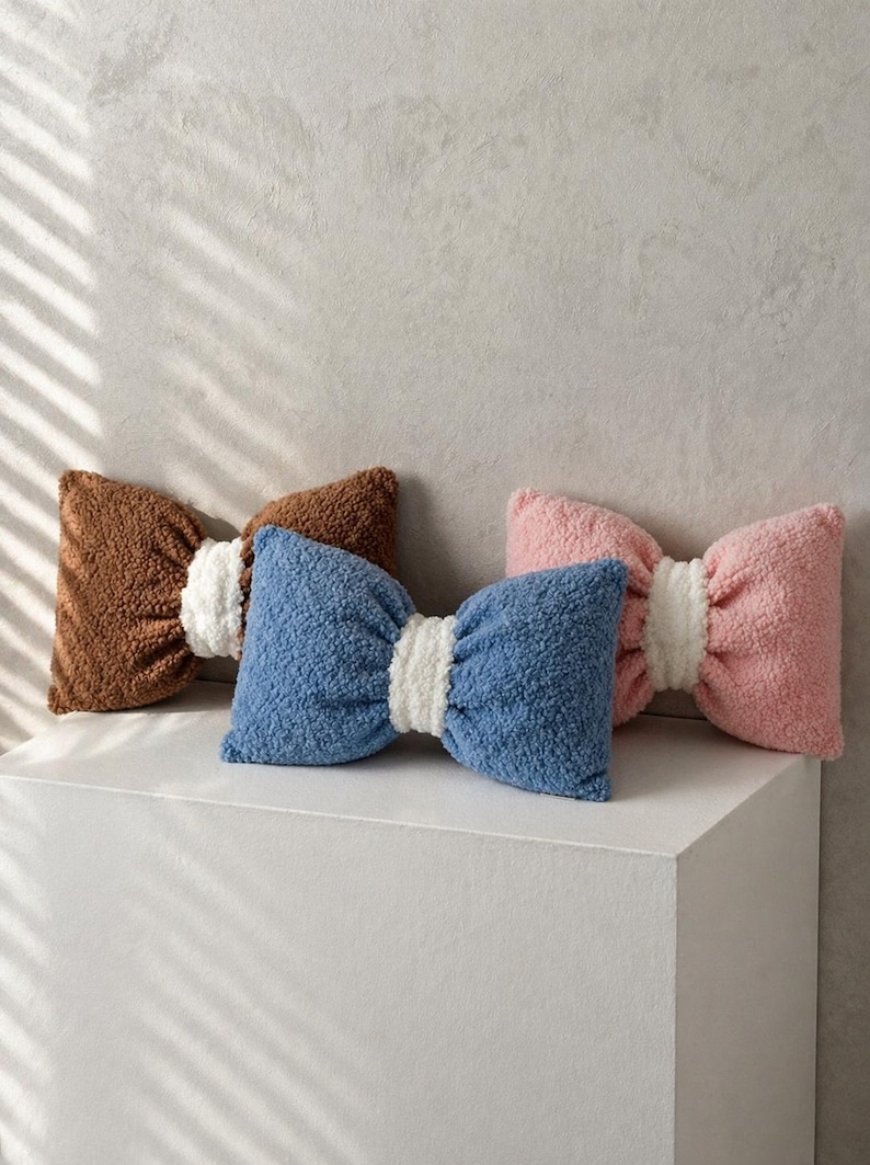 May include: Three bow-shaped decorative pillows in brown, blue, and pink. Each pillow has a white center and a textured, plush appearance. The pillows are arranged on a white surface against a neutral background.