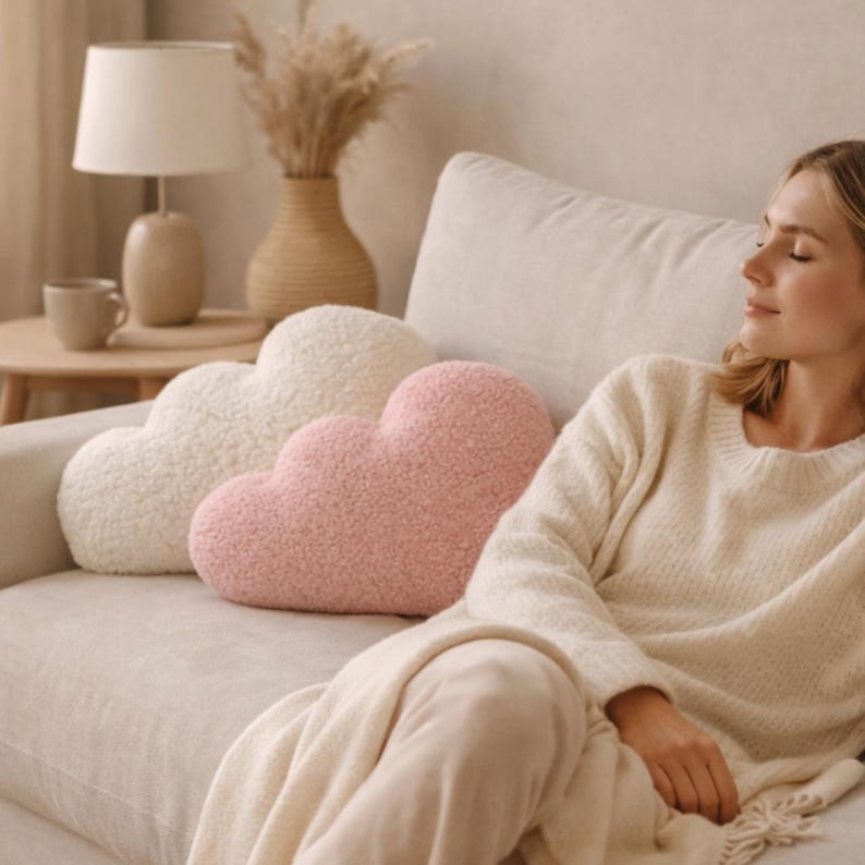 May include: Two cloud-shaped throw pillows in cream and pink rest on a beige sofa. The pillows have a soft, textured appearance. A woman wearing a cream-colored sweater is seated on the sofa.