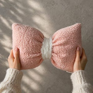 May include: A pink bow-shaped pillow with a white center. The pillow has a soft, textured appearance. The pillow is held by two hands wearing a cream-colored sweater. The background is a neutral gray.
