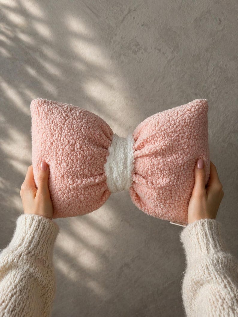 May include: A pink bow-shaped pillow with a white center. The pillow has a soft, textured appearance. The pillow is held by two hands wearing a cream-colored sweater. The background is a neutral gray.