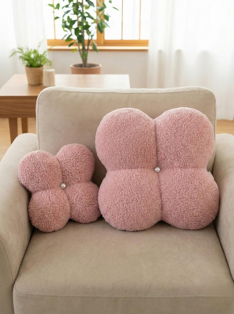 May include: Two pink, four-lobed decorative cushions with a textured, plush surface. Each cushion has a pearl-like button in the centre. The cushions are placed on a beige sofa, creating a cosy and inviting aesthetic.