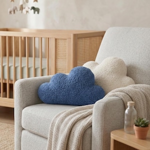 May include: A cozy nursery scene featuring a light gray armchair with two cloud-shaped pillows, one blue and one cream-colored. A beige blanket is draped over the arm. A wooden crib and mobile are visible in the background, creating a soft and inviting atmosphere.