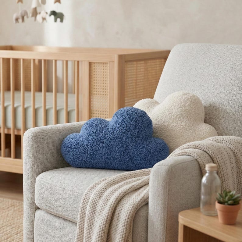 May include: A cozy nursery scene featuring a light gray armchair with two cloud-shaped pillows, one blue and one cream-colored. A beige blanket is draped over the arm. A wooden crib and mobile are visible in the background, creating a soft and inviting atmosphere.