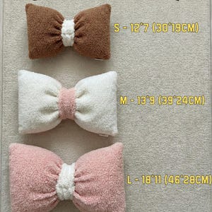 May include: Image shows a size chart for bow-shaped pillows. The chart displays three pillows in brown, white, and pink, with white centers. Sizes are S (12"x7"), M (13"x9"), and L (18"x11"). The text "SIZE CHART" is at the top.