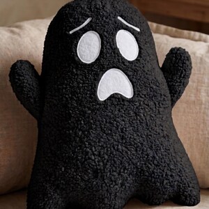 Pode incluir: Uma almofada de pel&uacute;cia em forma de fantasma preto, feita de tecido texturizado e encaracolado. A almofada apresenta dois olhos ovais brancos e uma boca branca, ambos com contornos costurados, criando uma express&atilde;o de surpresa com duas sobrancelhas.