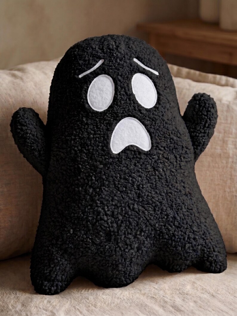 Pode incluir: Uma almofada de pel&uacute;cia em forma de fantasma preto, feita de tecido texturizado e encaracolado. A almofada apresenta dois olhos ovais brancos e uma boca branca, ambos com contornos costurados, criando uma express&atilde;o de surpresa com duas sobrancelhas.
