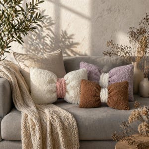 May include: A cozy living room scene features a gray sofa adorned with bow-shaped pillows in cream, brown, and lavender. A cream-colored throw blanket and a neutral-toned pillow add to the inviting ambiance. Decorative vases and greenery complete the warm, inviting setting.
