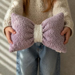 May include: A large, plush bow-shaped pillow in lavender and white. The pillow has a soft, textured appearance and is held by a person wearing a cream-colored sweater and blue jeans. The pillow is approximately 20 inches wide.