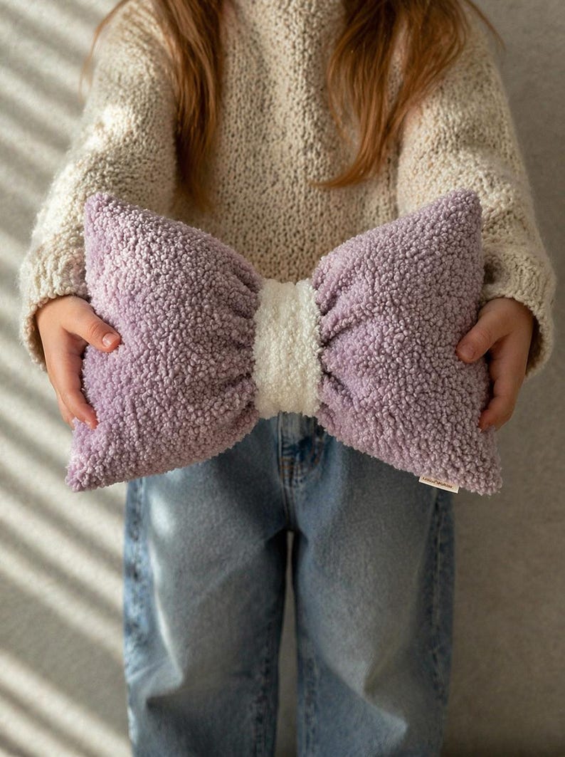 May include: A large, plush bow-shaped pillow in lavender and white. The pillow has a soft, textured appearance and is held by a person wearing a cream-colored sweater and blue jeans. The pillow is approximately 20 inches wide.